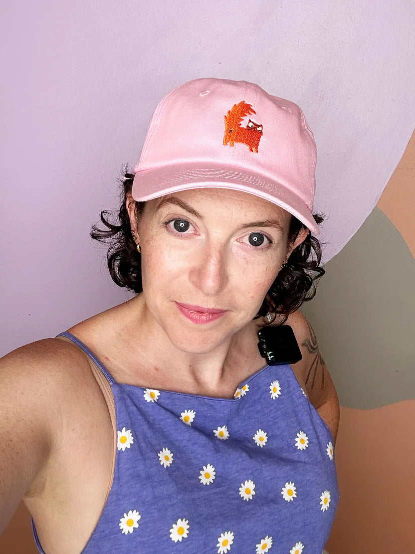 Person wearing a pink cap with an orange cat embroidered and a blue sleeveless top with white patterns against a plain background
