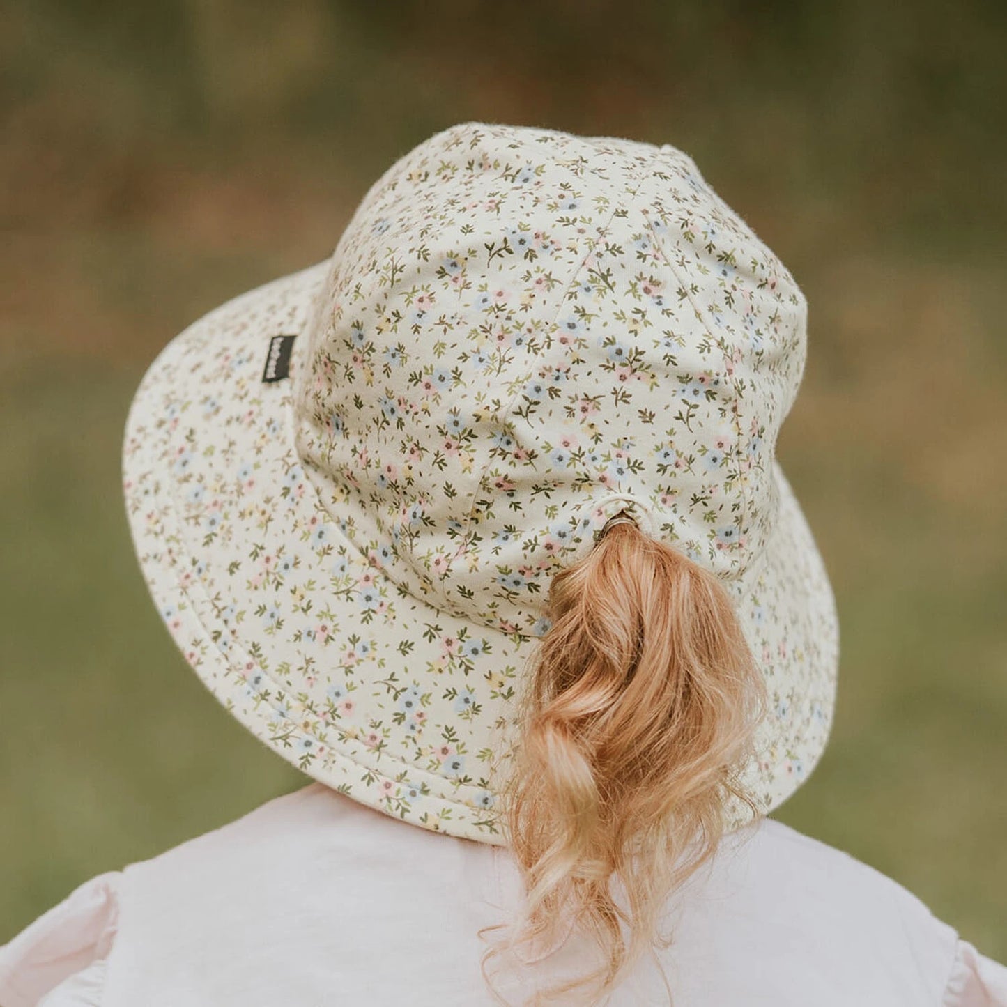 Child wearing a floral bucket hat with a ponytail slit in the back of hat and a blurred natural background