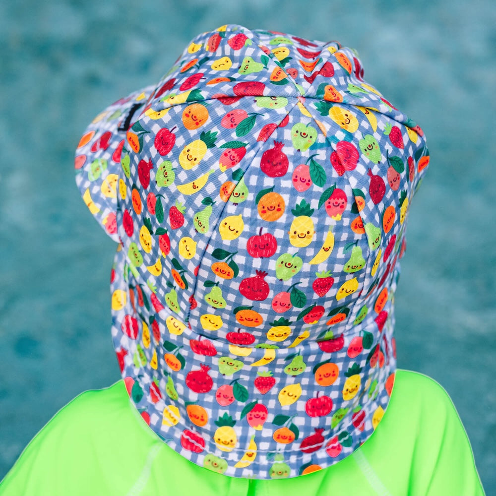 A back view of a child wearing a swim hat  featuring a blue and white gingham pattern and a colourful print of smiling cartoon fruits, including pineapples, strawberries and oranges. 