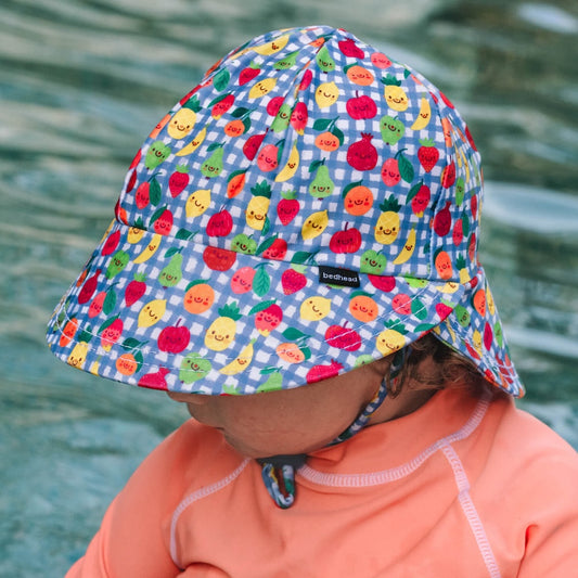 A child wearing a swim hat with a chin strap, featuring a blue and white gingham pattern and a colourful print of smiling cartoon fruits, including pineapples, strawberries and oranges. 