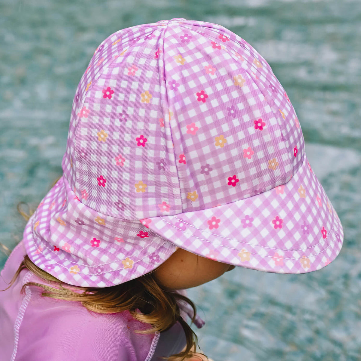 A child wearing a pink and white gingham swim hat with small floral prints, photographed outdoors with a pool in the background.
