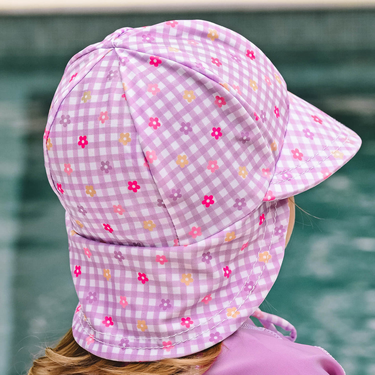 A child wearing a lavender and white gingham swim hat with small floral prints, photographed outdoors with a pool in the background.