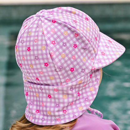 A child wearing a lavender and white gingham swim hat with small floral prints, photographed outdoors with a pool in the background.
