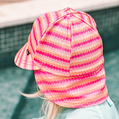 A close-up back view of a child wearing a pink and orange scalloped striped swim hat with a neck flap, set against a blurred pool background.