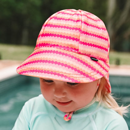 A child wearing a pink and orange scalloped striped swim hat with a light blue rashie, photographed outdoors near a pool.