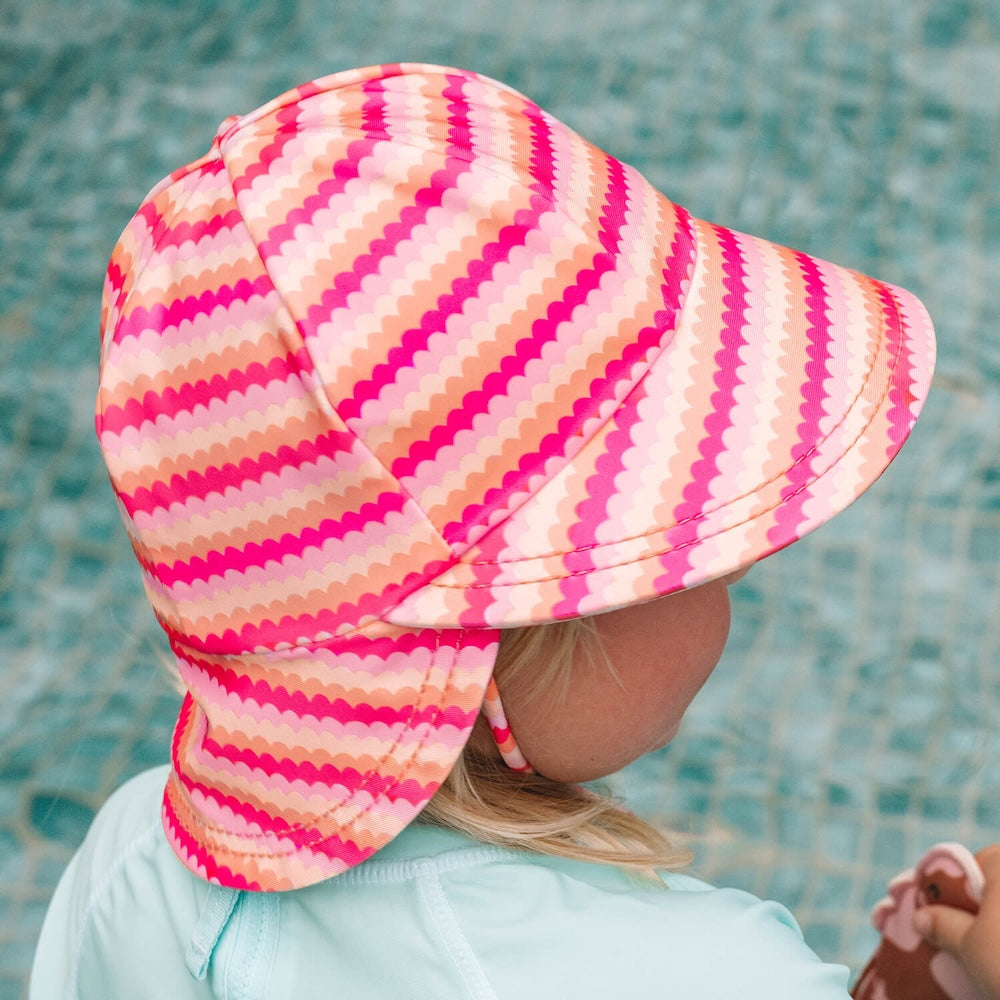 A child wearing a pink, orange and peach scalloped striped swim hat with a neck flap, photographed from behind against a tiled pool background.