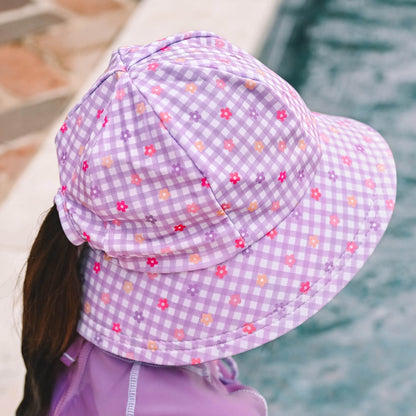A child wearing a lavender and white gingham swim bucket hat with small pink, purple and orange floral prints, photographed outdoors poolside.