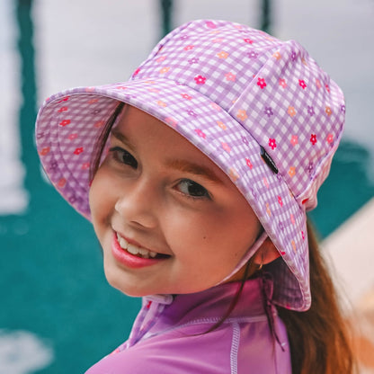 A young girl smiling and looking over her shoulder poolside, wearing a light purple and white gingham swim bucket hat with small multicolour floral prints and a purple rashie.