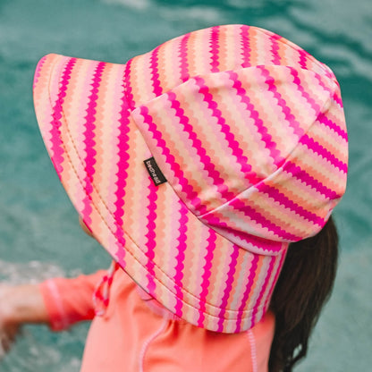 A child wearing a swim bucket hat with a pink, orange and cream scalloped stripe pattern, photographed outdoors with water in the background.