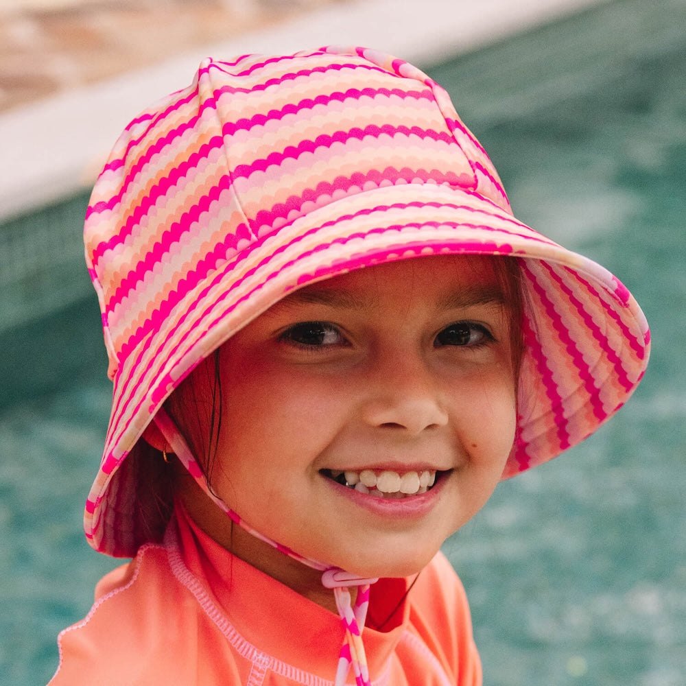 A young child wearing a pink and peach scalloped striped swim bucket hat with a chin strap, standing by a swimming pool.