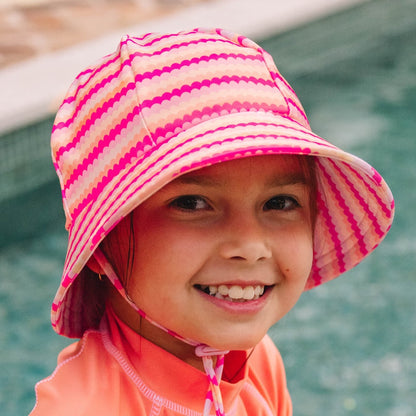 A young child wearing a pink and peach scalloped striped swim bucket hat with a chin strap, standing by a swimming pool.