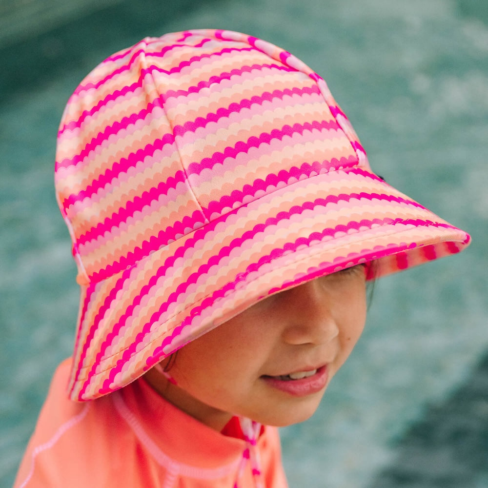 A close-up of a child wearing a pink and peach striped swim bucket hat with a scalloped stripe pattern, paired with a coral orange rashie, set against a pool in the background.