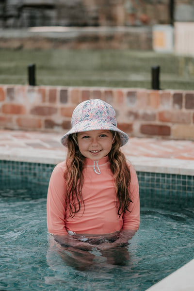 front view of Bedhead Blossom Swim Bucket Hat worn by a young model wearing a coral coloured rashy and standing in a swimming pool