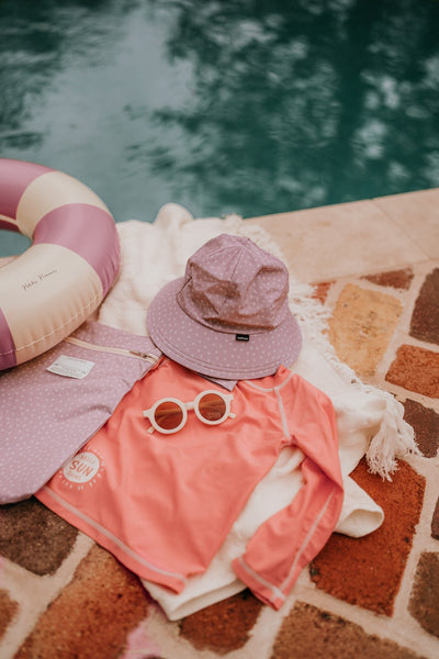 flat lay of Bedheket Hat with a pair of sunnies, a rashy, a bag, a towel, a floatation device next to a swimming poolad Lila Swim Buc