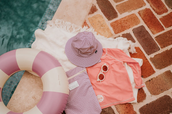 flat lay of Bedhead Lila Swim Bucket Hat with a pair of sunnies, a rashy, a bag, a towel, and a floatation device next to a swimming pool