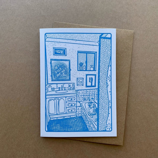 A greeting card featuring a blue line-art illustration of a bathroom interior with the mirror above the sink reading "hey good looking" and a border collie in the bathtub, placed on top of a brown kraft envelope against a brown kraft paper background.