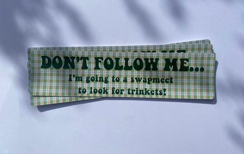 Carla Adams Swapmeet Bumper Sticker. The sticker is rectangular with a green and light brown plaid pattern. The black text reads "DON'T FOLLOW ME.. I'm going to a swapmeet to look for trinkets!".