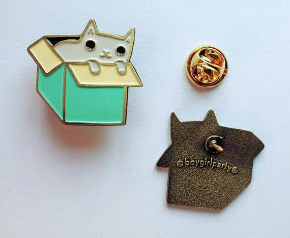 A grey cat enamel pin displayed on a white background. The front of the pin shows a grey cat peeking out of a mint green box with gold-tone metal outlines.