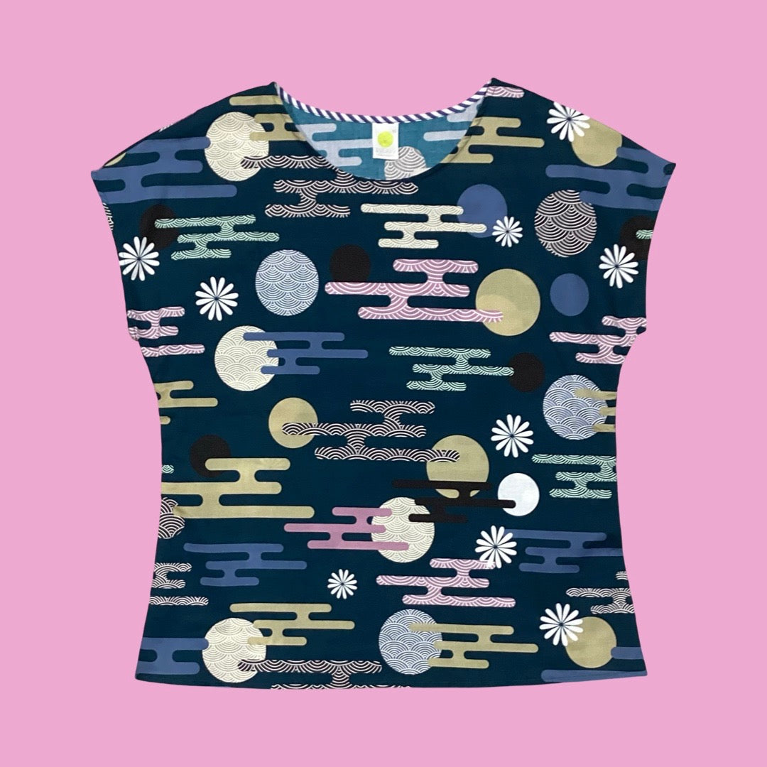 Essaye Tille short-sleeved top with a dark teal base, featuring a colourful abstract pattern of circles, wavy lines, and floral motifs in pastel tones, laid flat against a soft pink background.
