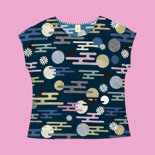 Essaye Tille short-sleeved top with a dark teal base, featuring a colourful abstract pattern of circles, wavy lines, and floral motifs in pastel tones, laid flat against a soft pink background.