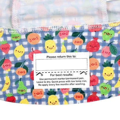 Close-up of the name label on the inside brim of a bucket hat, featuring a colorful print of smiling fruits like pineapples, apples and oranges on a blue gingham background.
