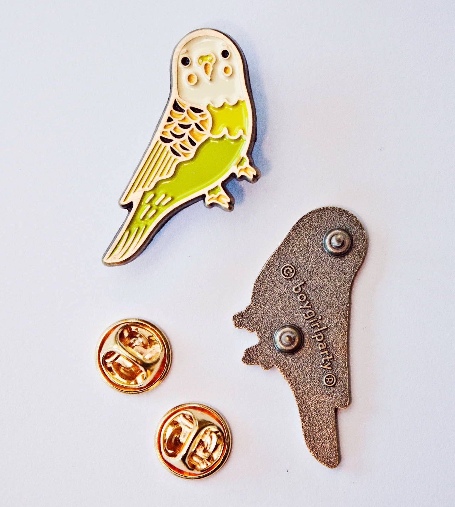 A bird-shaped enamel pin displayed on a white background. The pin is gold-toned with light green, cream, and black detailing, shown alongside two gold-toned pin backs and a metal backing piece.