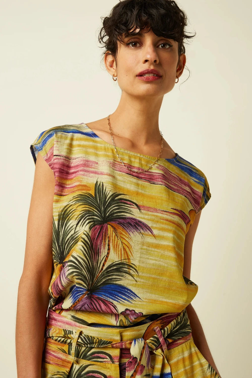 Woman wearing a colourful sleeveless top with a vintage-inspired tropical palm tree print on a plain beige background