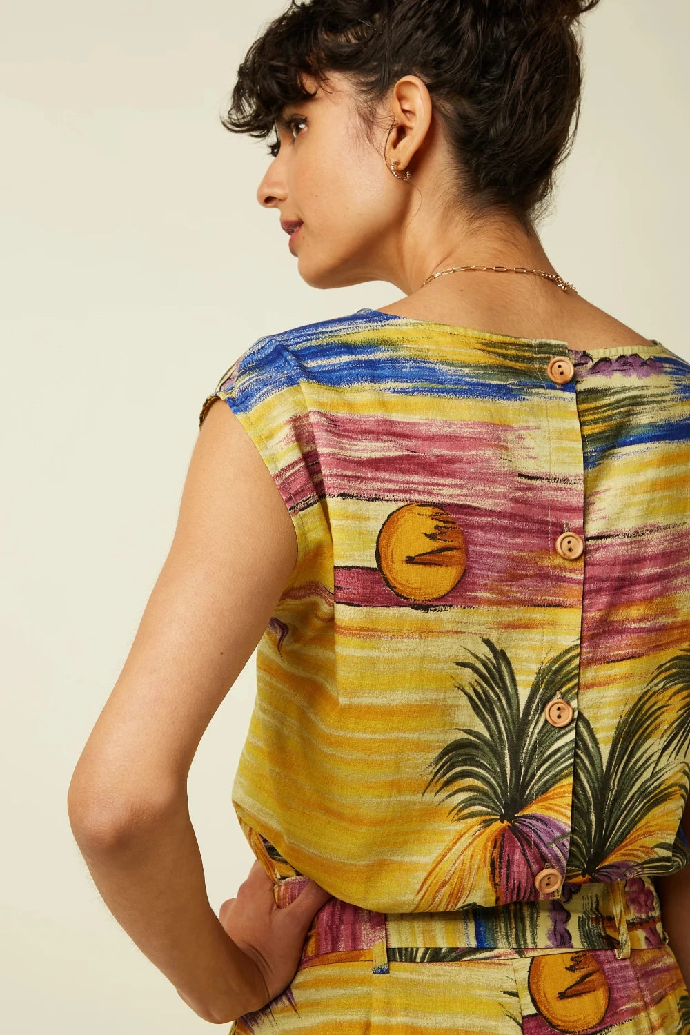A woman shown from the back wearing a sleeveless top with a colourful, vintage-inspired tropical sunset print featuring yellow, pink, blue, and green palm tree details, set against a plain beige background