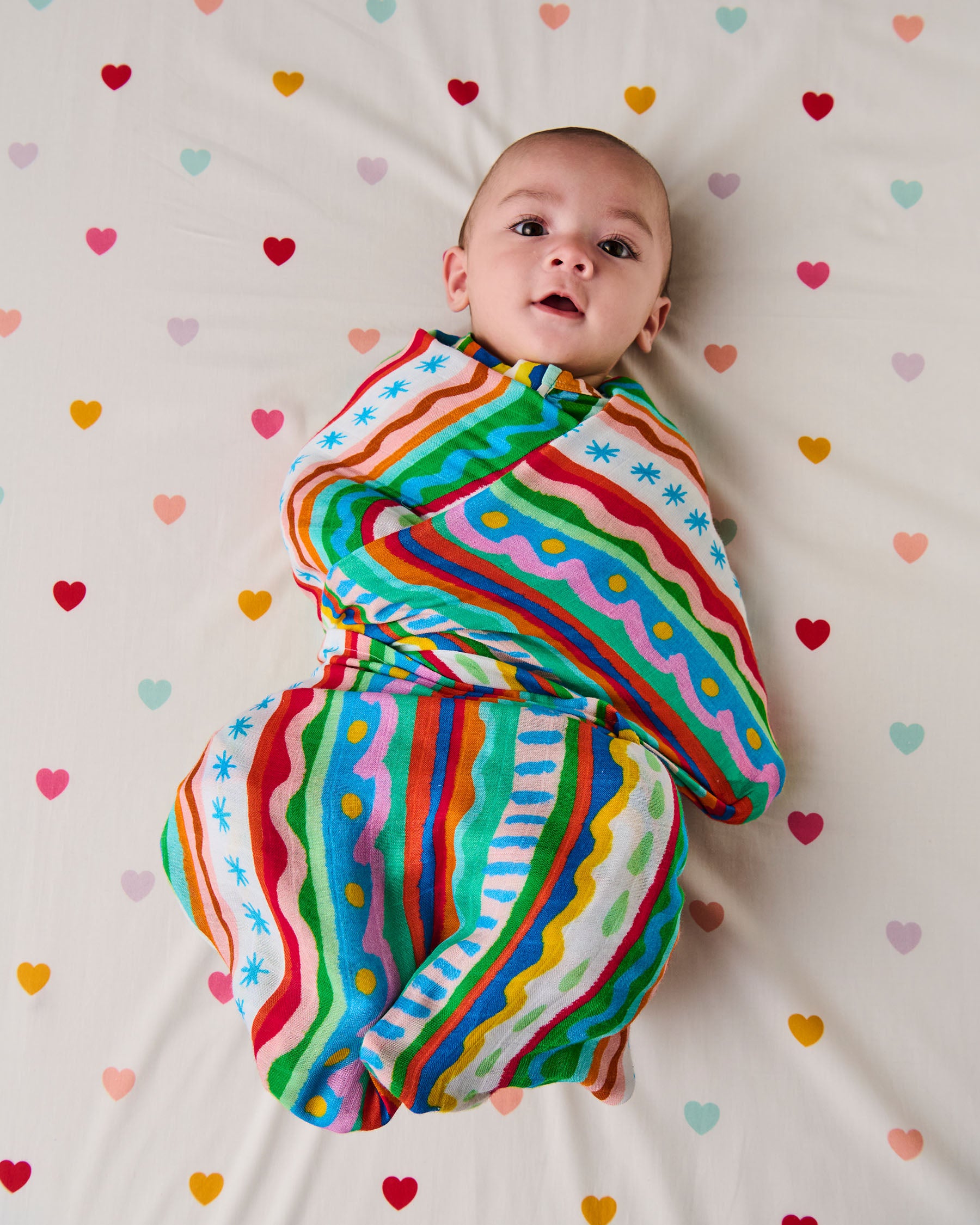 Baby wrapped in a colorful striped swaddle on a heart-patterned blanket