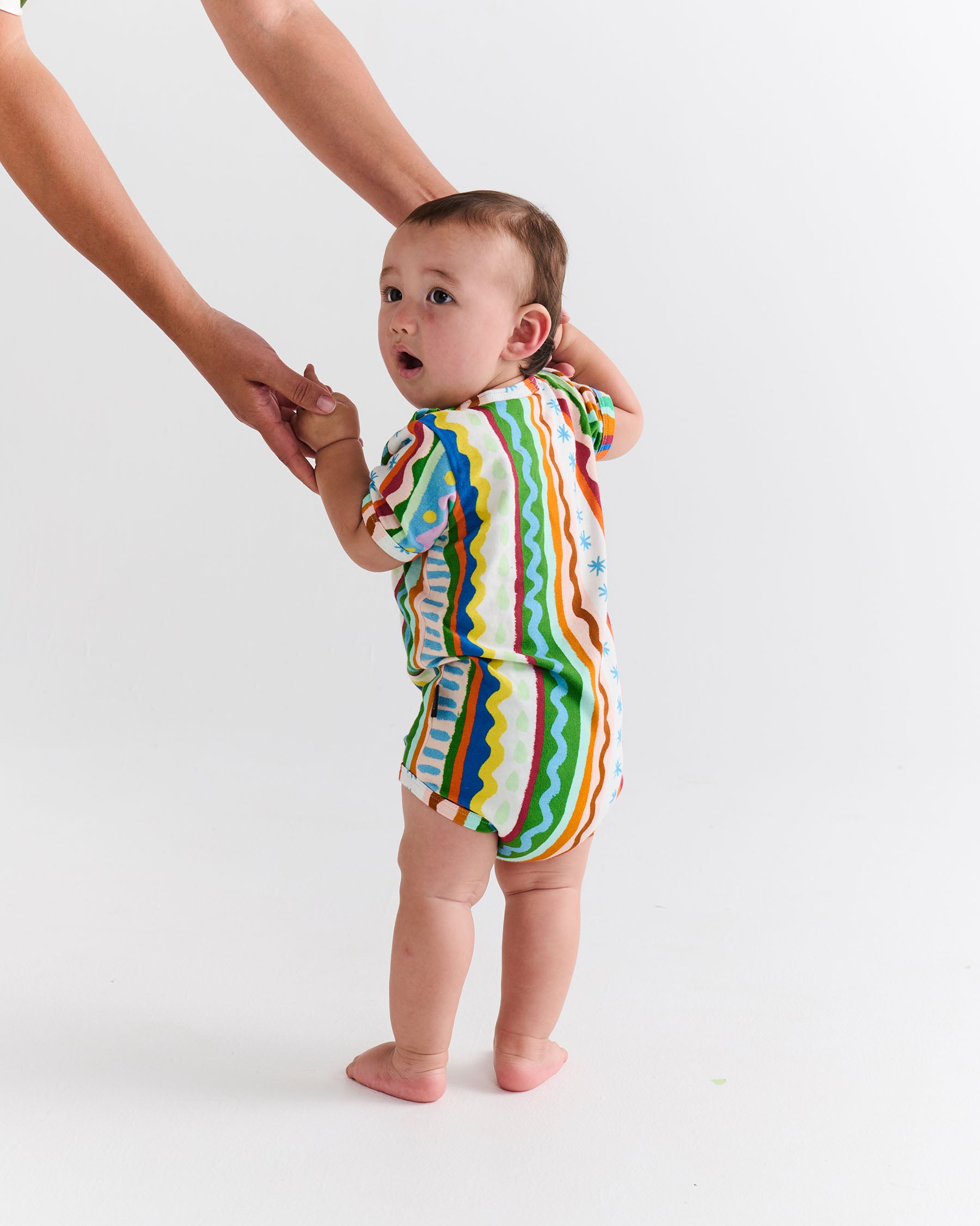 Baby in a colorful striped onesie standing with assistance on a white background