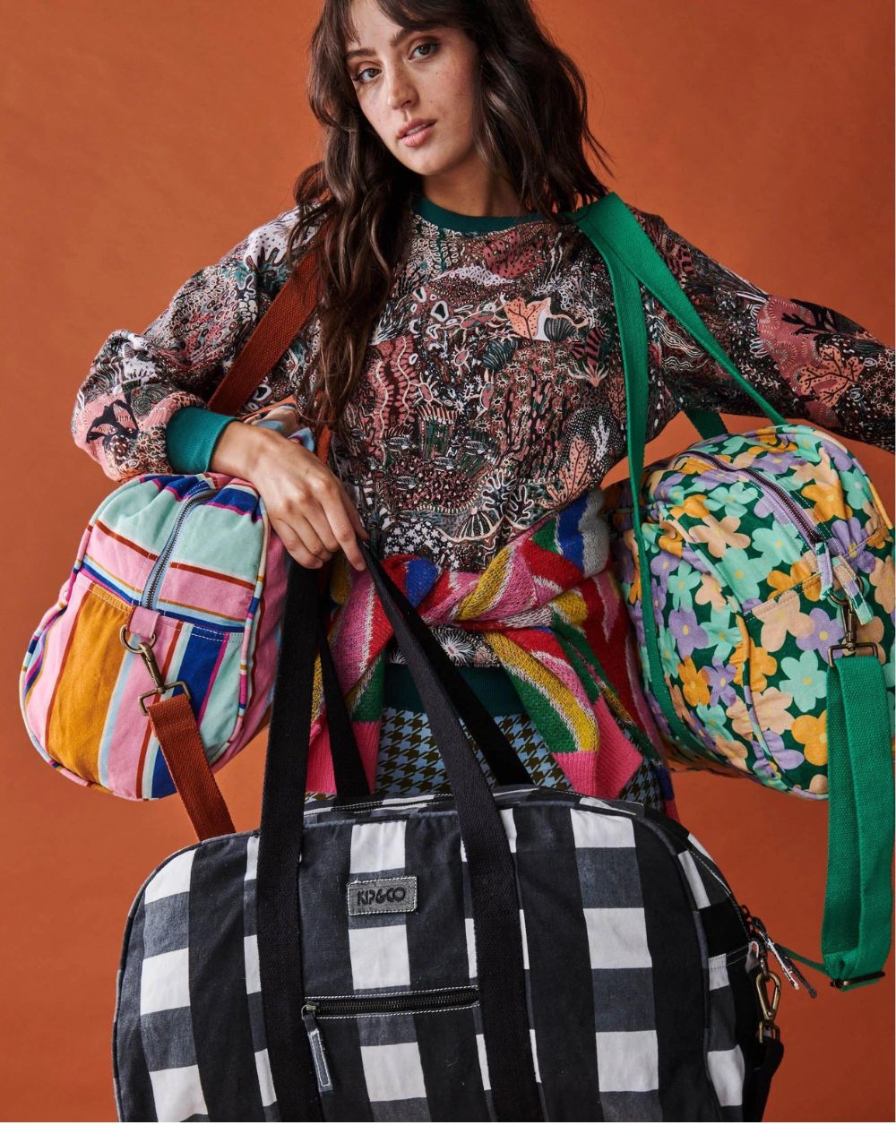 Model is holding Kip & Co Black & White Gingham Duffle Bag in her hand, and carrying 2 other duffle bags of different designs on her shoulders.