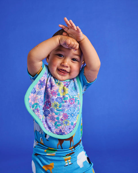 Styled image of a smiling young toddler standing with their hands above their head and wearing the Kip & Co Bunch of Fun Bib.