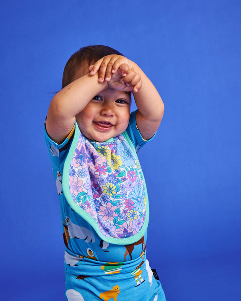 Styled image of a young toddler standing with their arms above their head and wearing the Kip & Co Bunch of Fun Bib.