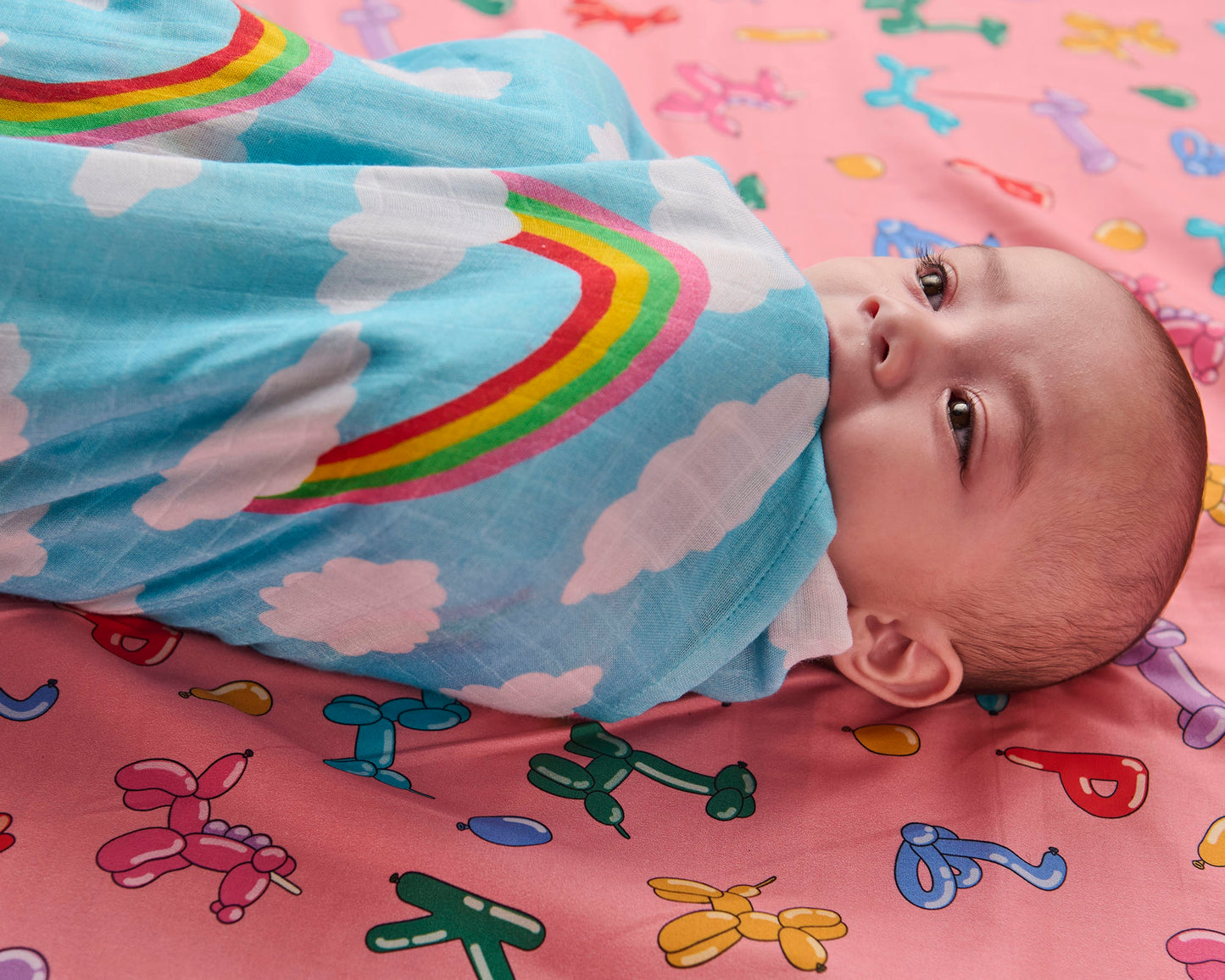 Baby wrapped in a colorful blanket with rainbows and clouds on a pink blanket with cartoon designs.