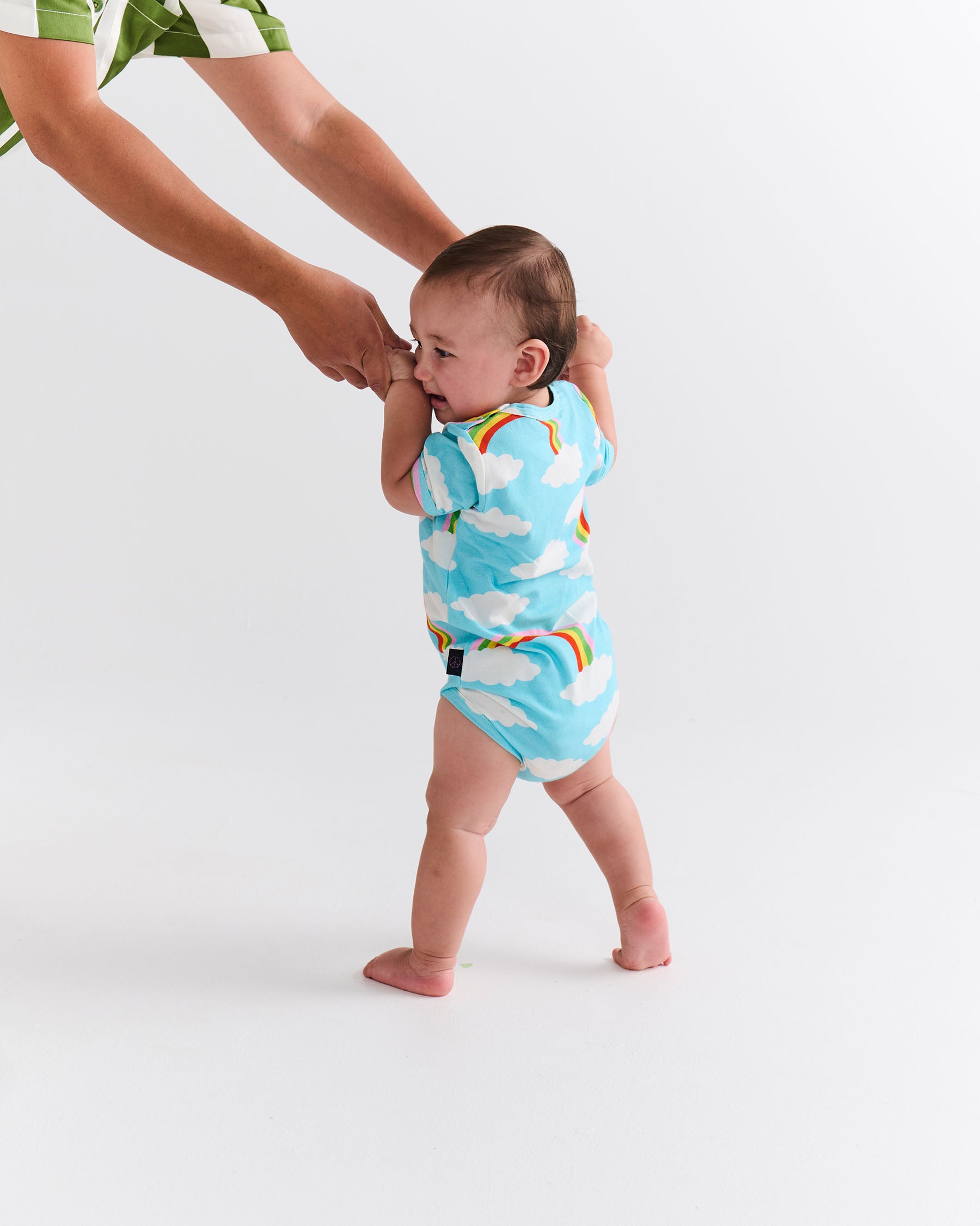 Baby in a colorful cloud and rainbow onesie being held by an adult on a white background