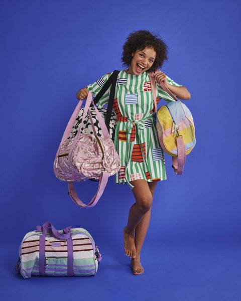 Model is posing with multiple Kip & Co duffle bags. She is holding the Kip & Co Palmy Duffle Bag, on her shoulders is a Kip & Co Corfu Tartan Duffle Bag, and on her other shoulder is a black and white duffle bag. On the floor next to her is the Kip & Co Island Delight Duffle Bag.