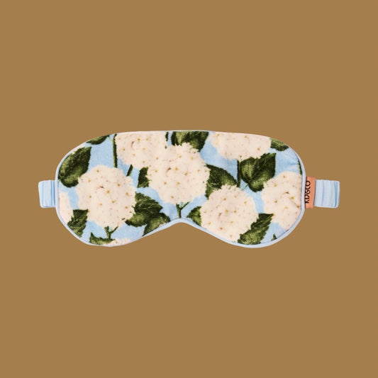 A plush sleep mask with a light blue background and a white flower and green leaf pattern, featuring light blue straps and a small orange brand tag that reads 'kip&co'. Background is a solid light brown.