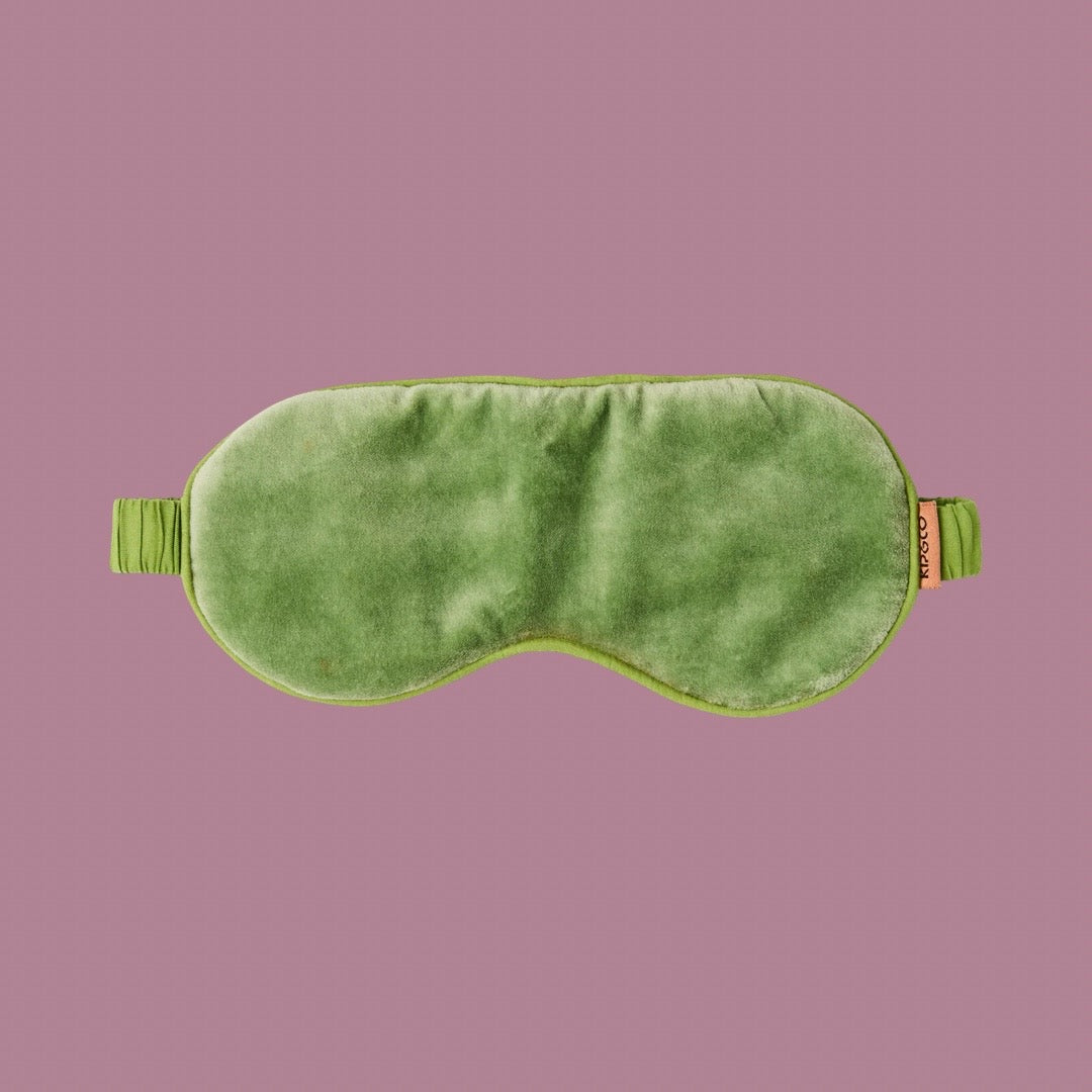 A pesto-green velvet sleeping mask with elasticated strap. The 'Kip&Co' label can be seen on the side of the mask. Light mauve-purple background.