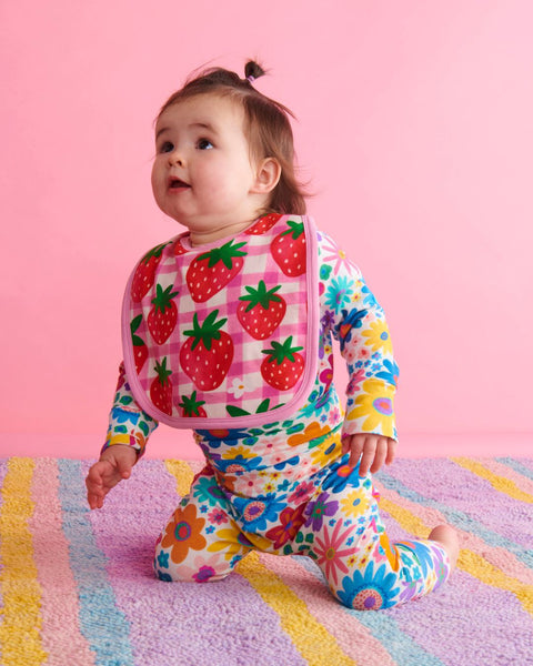 A toddler wearing the Kip & Co Strawberry Jam Bib and a onesies, kneeling on the floor looking upwards.