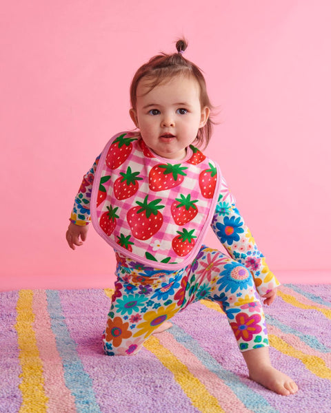 A toddler wearing the Kip & Co Strawberry Jam Bib and a onesies, kneeling on the floor.