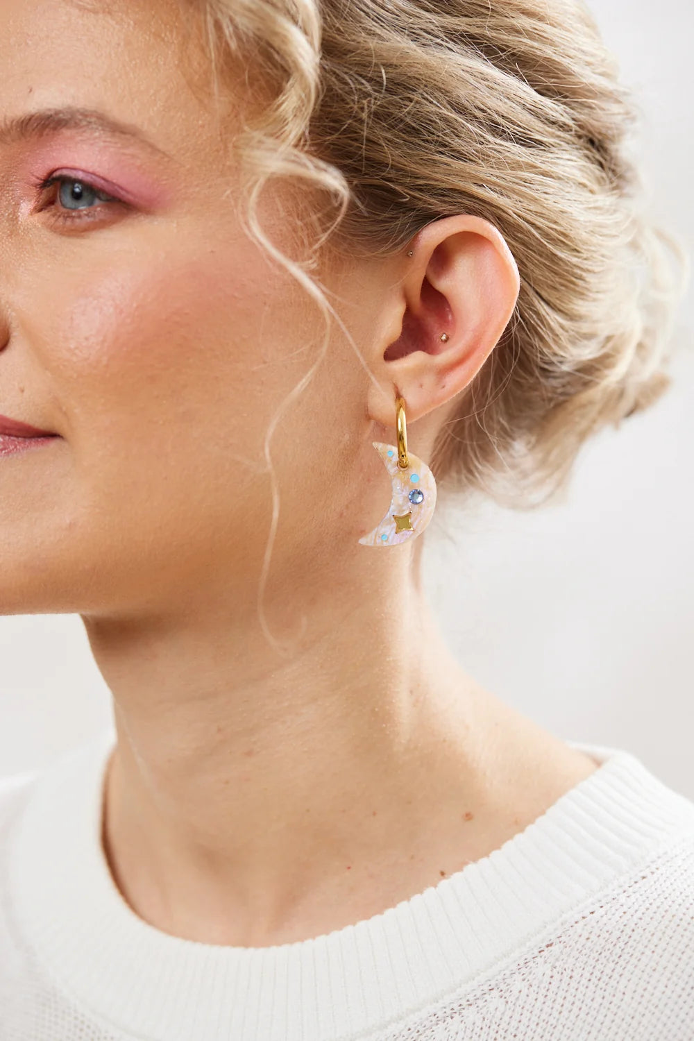 A close-up of a woman wearing a gold crescent moon earring with two small stones in the centre against a soft light background.