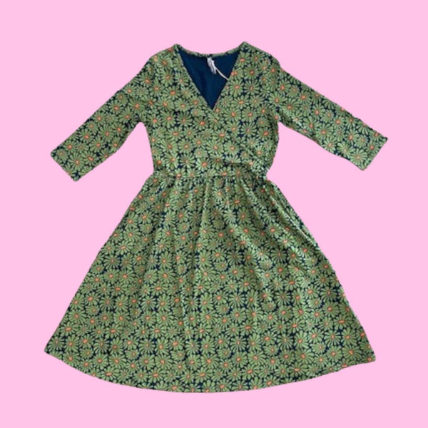 Mata Traders Callie Wrap Dress in Olive Floral. This dress has a pattern full of olive coloured daisy-like flowers with a bright orange centre, and a navy blue background.