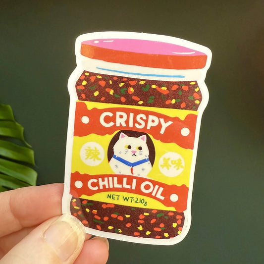 Sticker by Naomi Wilkinson of a jar labelled 'Crispy Chilli Oil' with a picture of a cat, held by a hand against a green background.