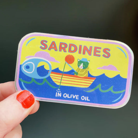 Colourful 'Sardines in Olive Oil' sticker held by a hand with red nail polish against a dark background.