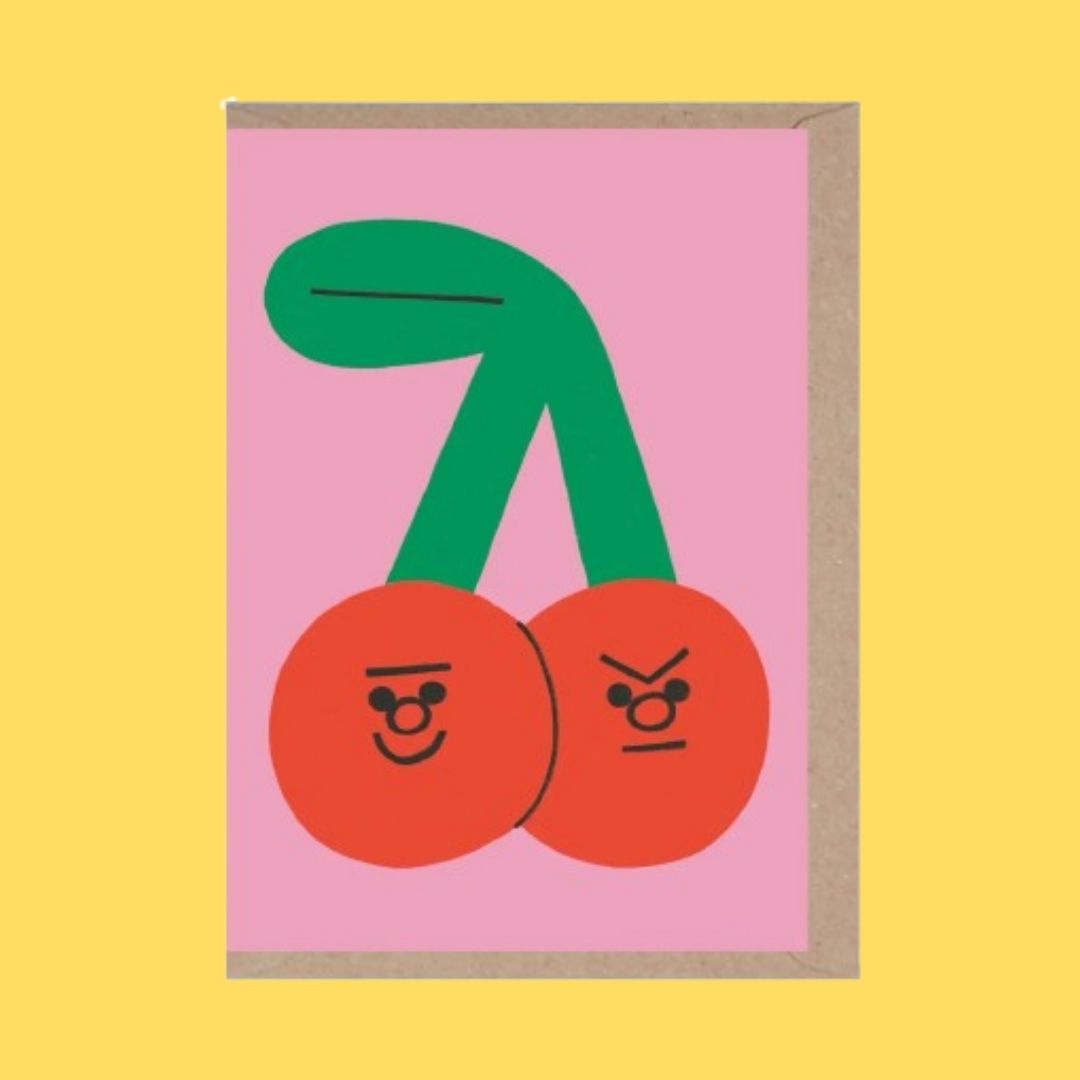 A greeting card with a pink background featuring a stylized illustration of two red cherries with green stems and simple facial expressions, shown with a kraft envelope on a solid yellow background.