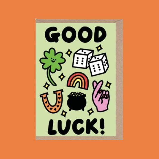 A Good Luck greeting card with a light green background and a brown kraft envelope, featuring cartoon illustrations of a four-leaf clover, dice, rainbow, horseshoe, pot of gold, and a pink luck hand, with the text 'GOOD LUCK!' in black.