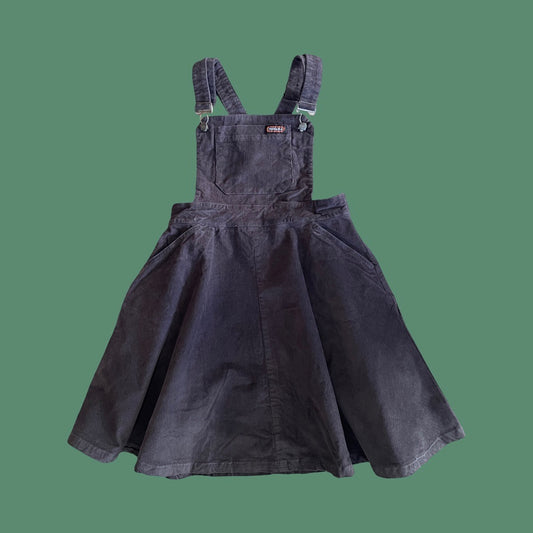 A Run & Fly charcoal corduroy pinafore dress with adjustable shoulder straps and a flared skirt, shown against a solid green background.