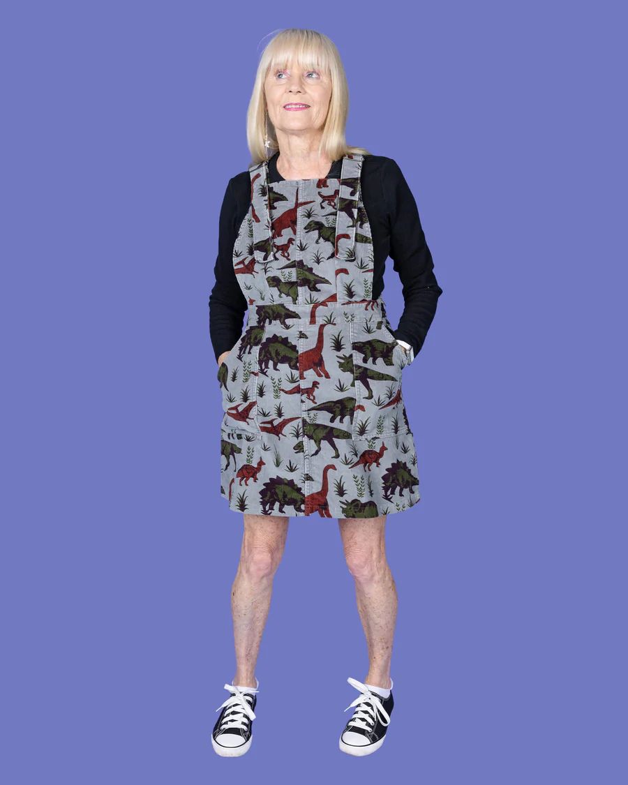 Model is wearing Run and Fly Dino Corduroy Pinafore. She is looking away from the camera with her hands in the pockets.