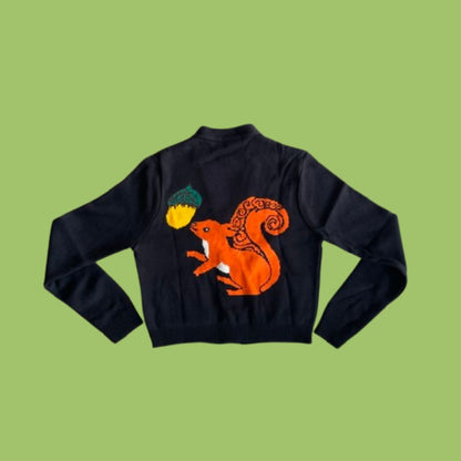 A black knit sweater with a large orange and yellow embroidered squirrel holding an acorn on the back, displayed flat against a solid light green background.