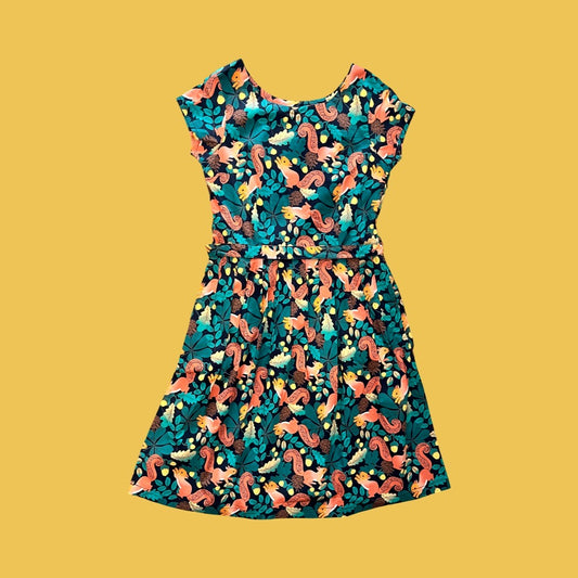A cap-sleeve knee-length vintage style dress with a colourful print featuring squirrels among autumn foliage and pine cones on a black base, displayed flat against a solid mustard yellow backdrop.